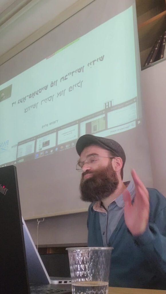 siedząca osoba wygłaszająca referat, w tle tekst w języku jidysz